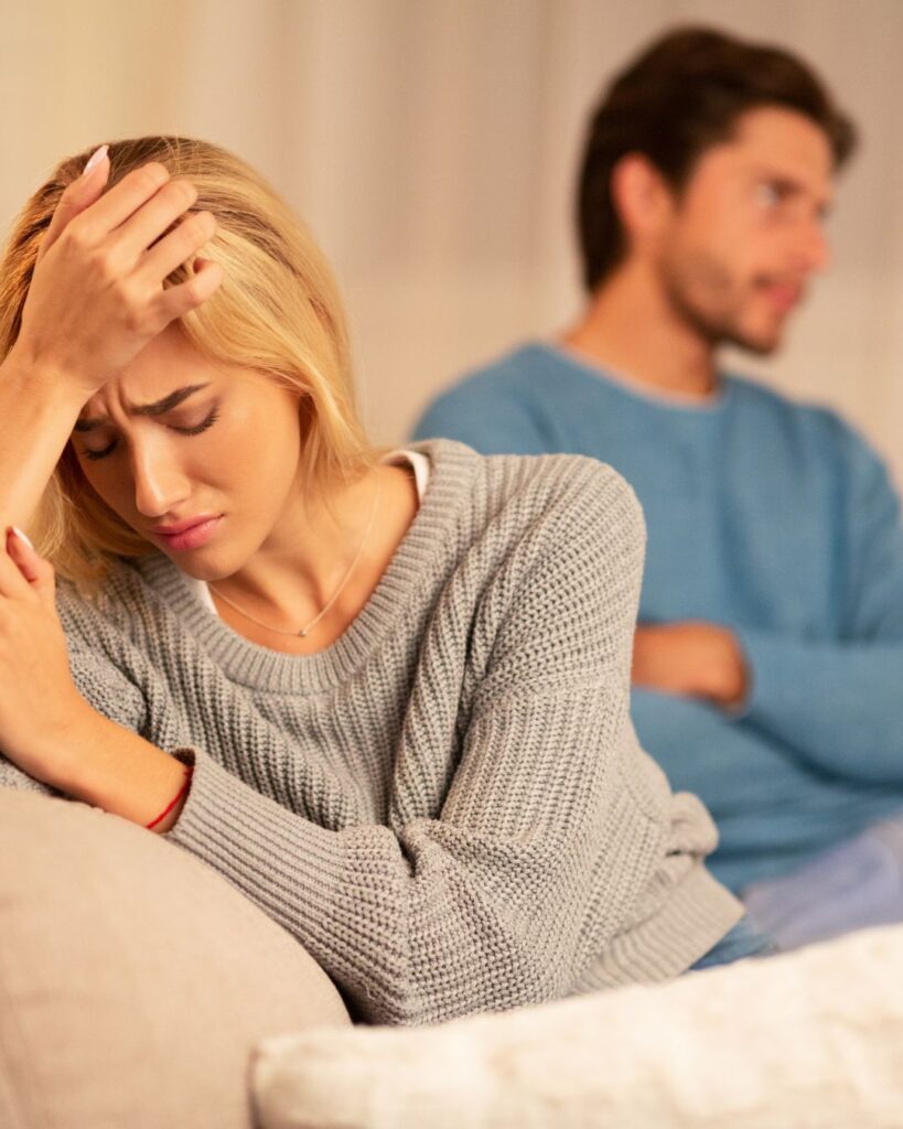 Couple sat on sofa, woman visibly upset after discovering partners betrayal, man sat with arms crossed.