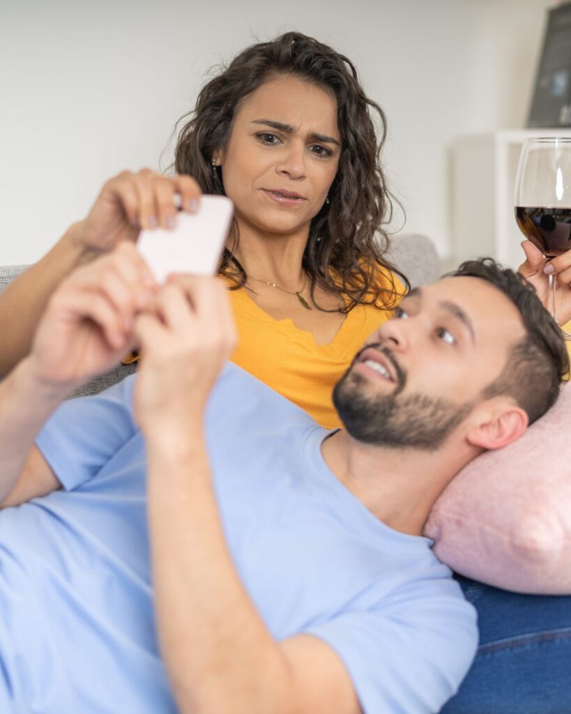 Man is laid down on sofa on his phone, his partner is reaching for the phone and looking at it with a worried look on her face