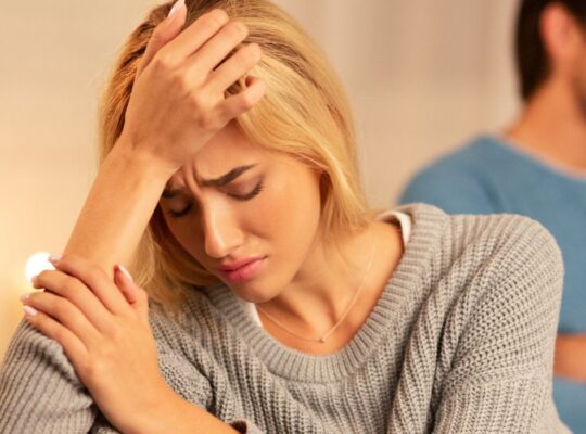 Couple on sofa, man with arms crossed looking annoyed, woman visibly distressed after discovering her partners betrayal