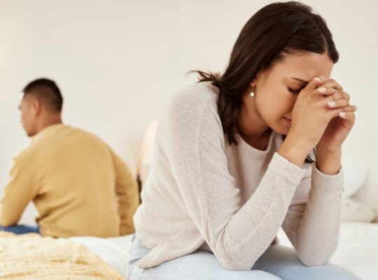 Woman upset sat on one side of bed, man sat on other side with his back to her after discovery of his betrayal